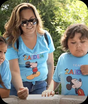Woman and two children wearing matching blue shirts with Mickey Mouse, outdoors with greenery in the background.
