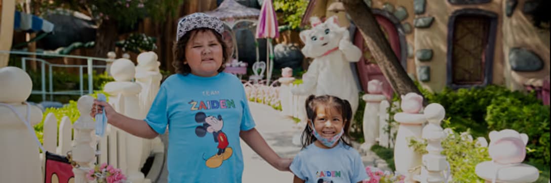 Two children holding hands and smiling, wearing matching blue Mickey Mouse shirts in a themed park setting