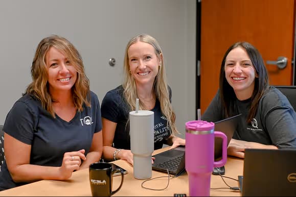 Three smiling women sitting at a table with laptops and coffee mugs in an office setting.