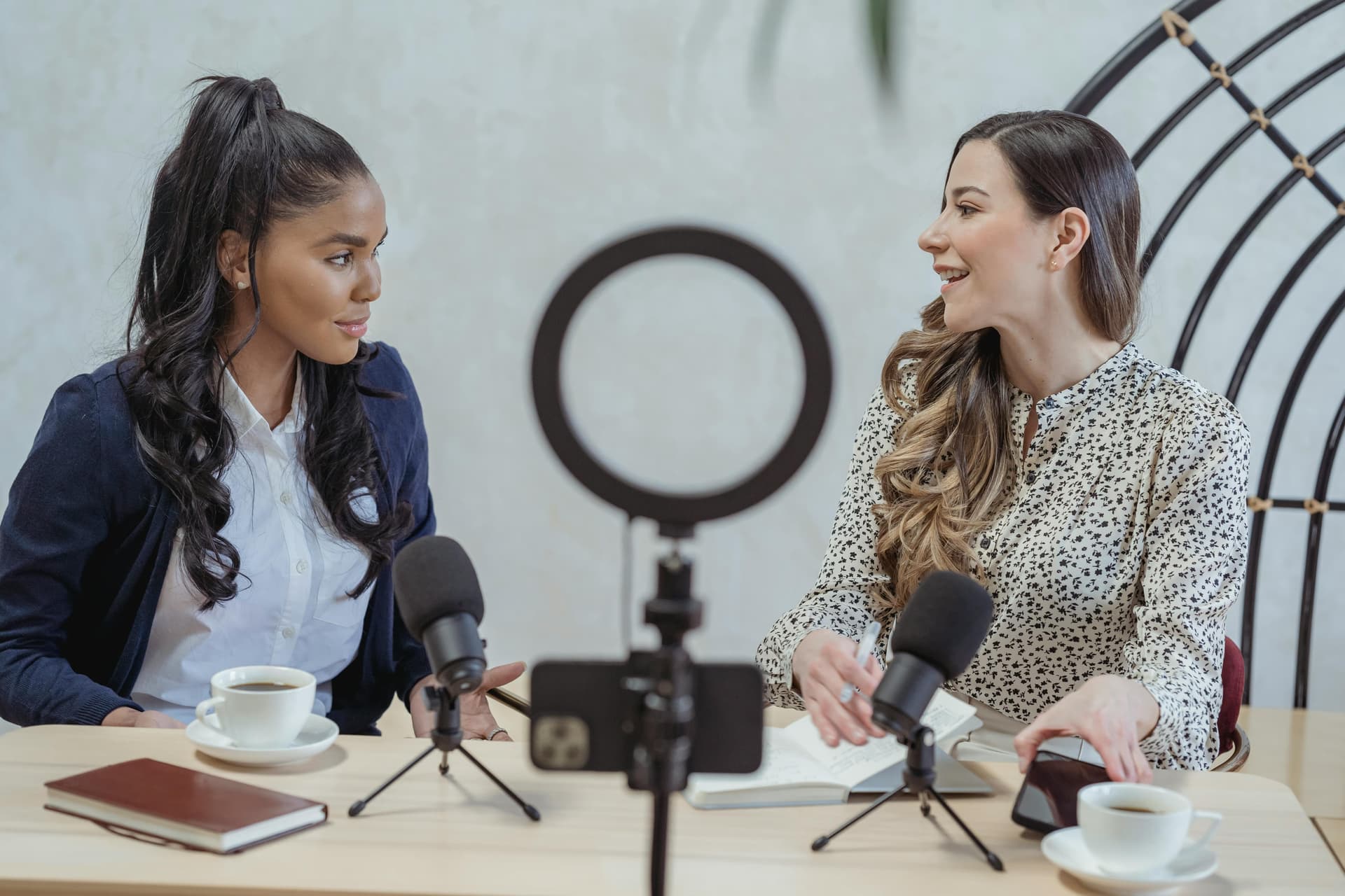 Two women sitting at a table, recording a podcast with microphones, a phone on a tripod, and cups of coffee.