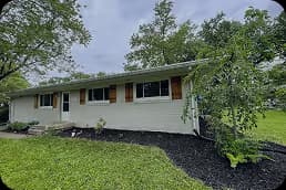 Single-story white brick house with brown shutters surrounded by green grass and trees under a cloudy sky