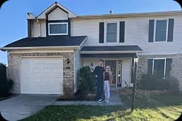 Two people standing on the front lawn in front of a two-story house with white siding and stone accents