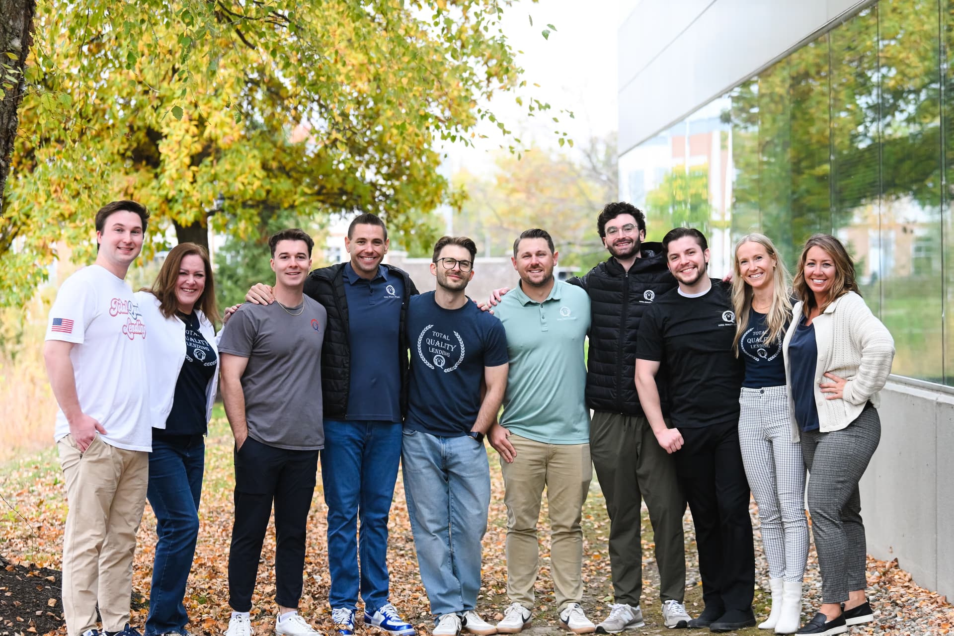 Group of ten people standing closely outdoors in front of a tree and a building with glass windows, smiling and casually dressed in autumn.