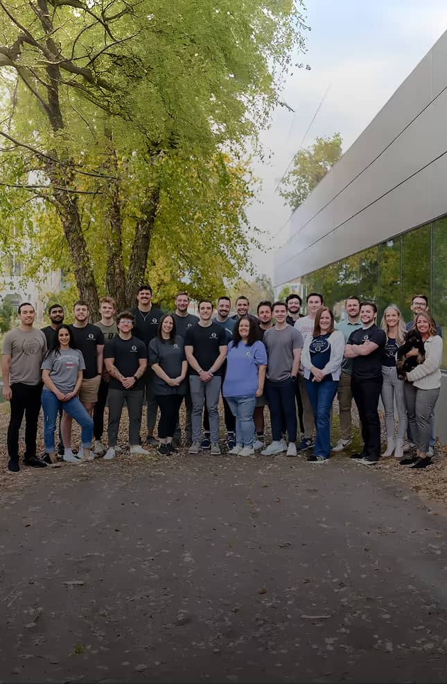 Group of diverse people standing outdoors in front of trees and a modern building, posing for a team photo.